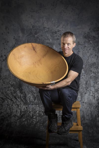 En man sitter på en pall och håller en mycket stor skål gjord av trä i sina händer.